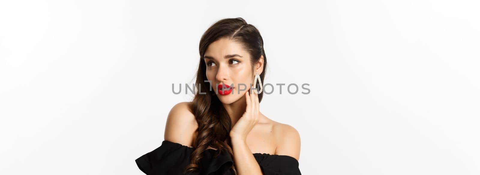 Fashion and beauty concept. Close-up of elegant woman in black dress, showing earrings and looking sensual, red lipstick and makeup on, white background.