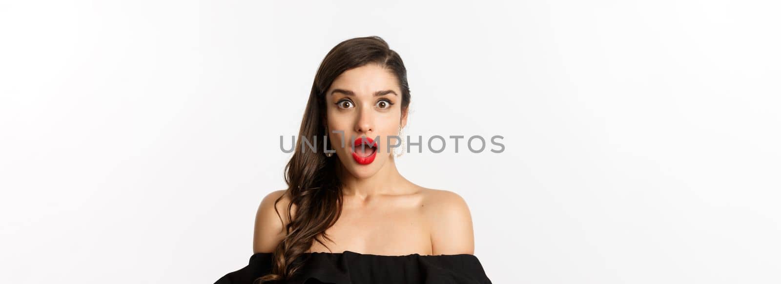 Fashion and beauty concept. Close-up of attractive brunette woman in black dress open mouth surprised, looking in awe at camera, white background.