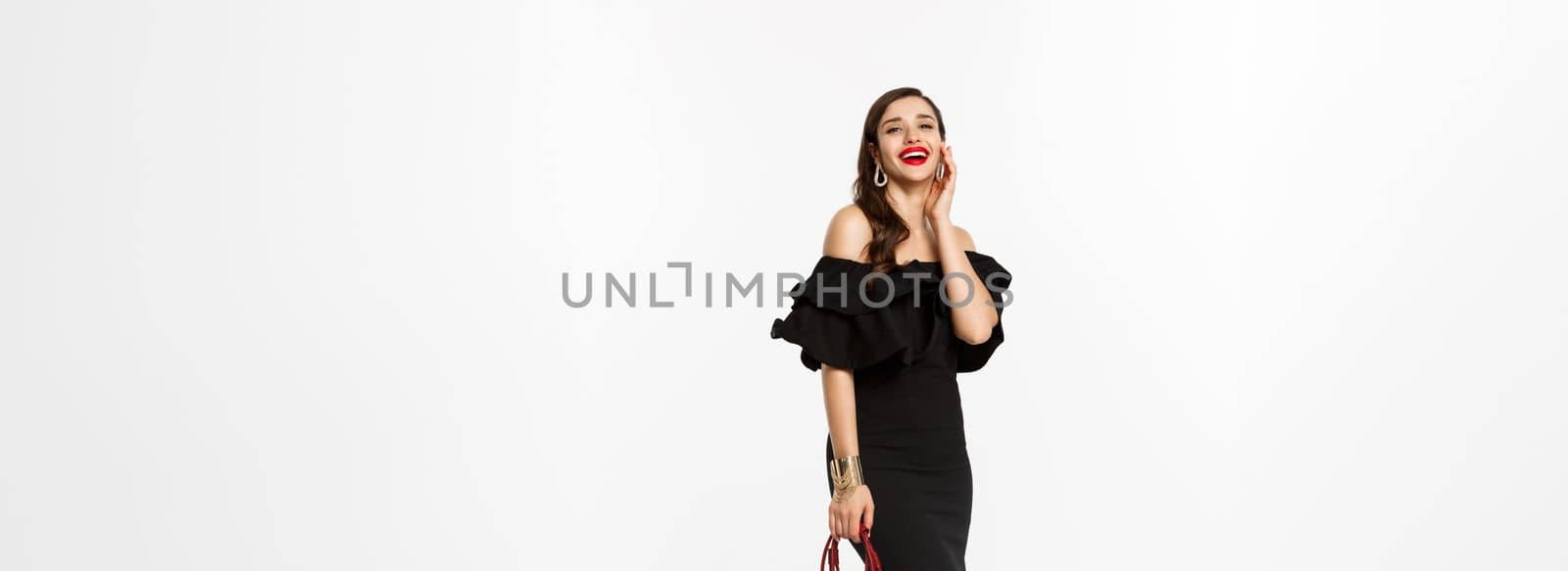 Beauty and fashion concept. Full length of beautiful young lady going on party in heels, black dress and makeup, holding elegant purse, laughing at camera, white background.