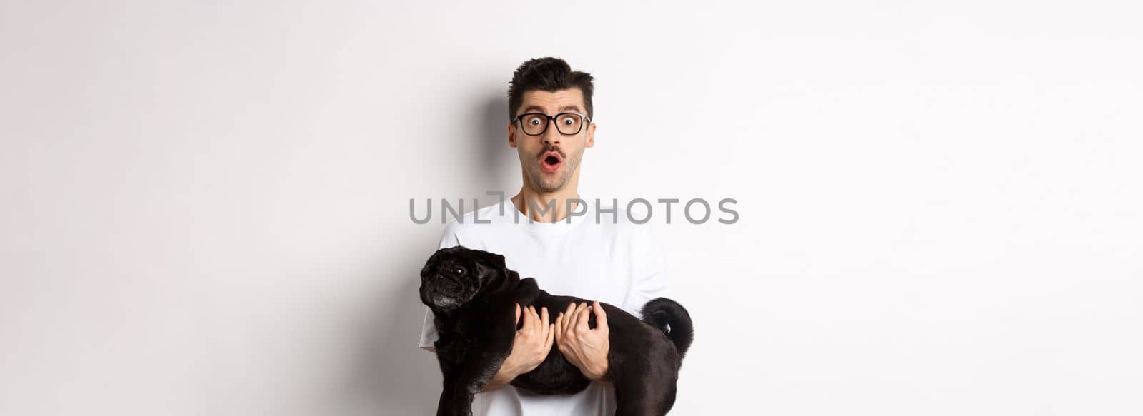 Surprised young man in glasses holding cute black pug, dog owner staring at camera with impressed face, saying wow, standing over white background.