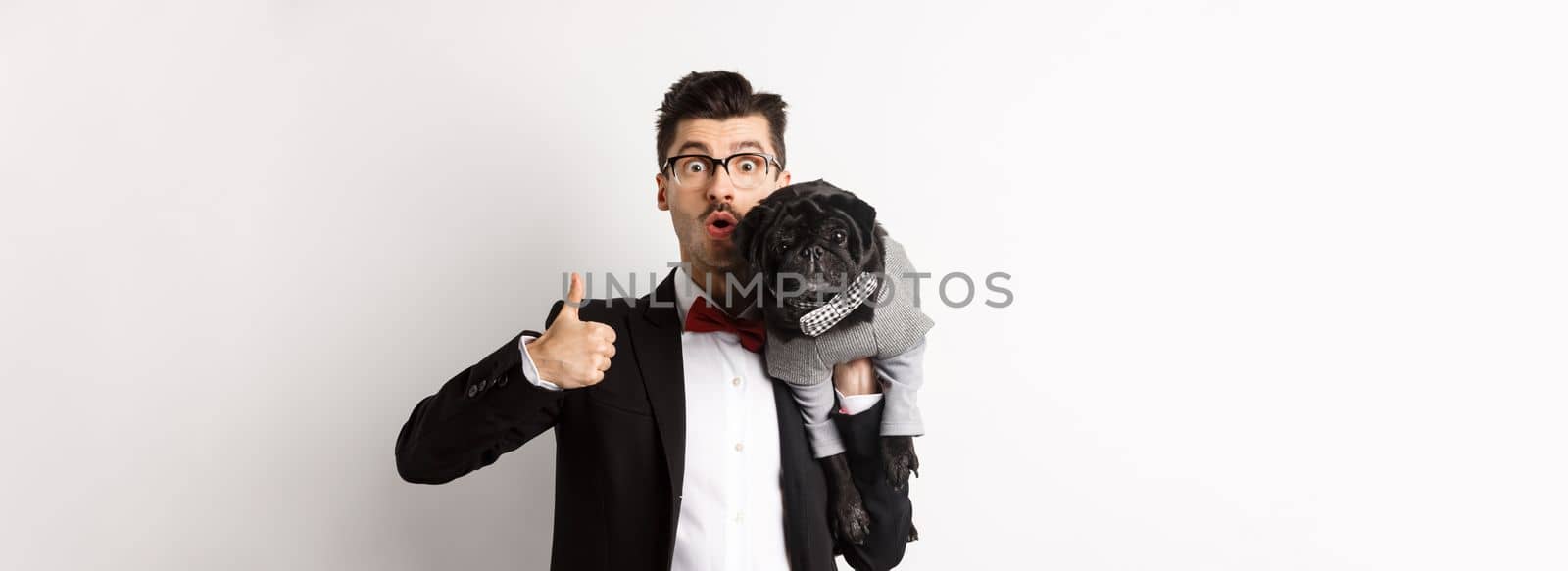 Happy young hipster in suit and glasses, showing thumb-up, holding cute black dog on shoulder, love his pug, standing over white background.