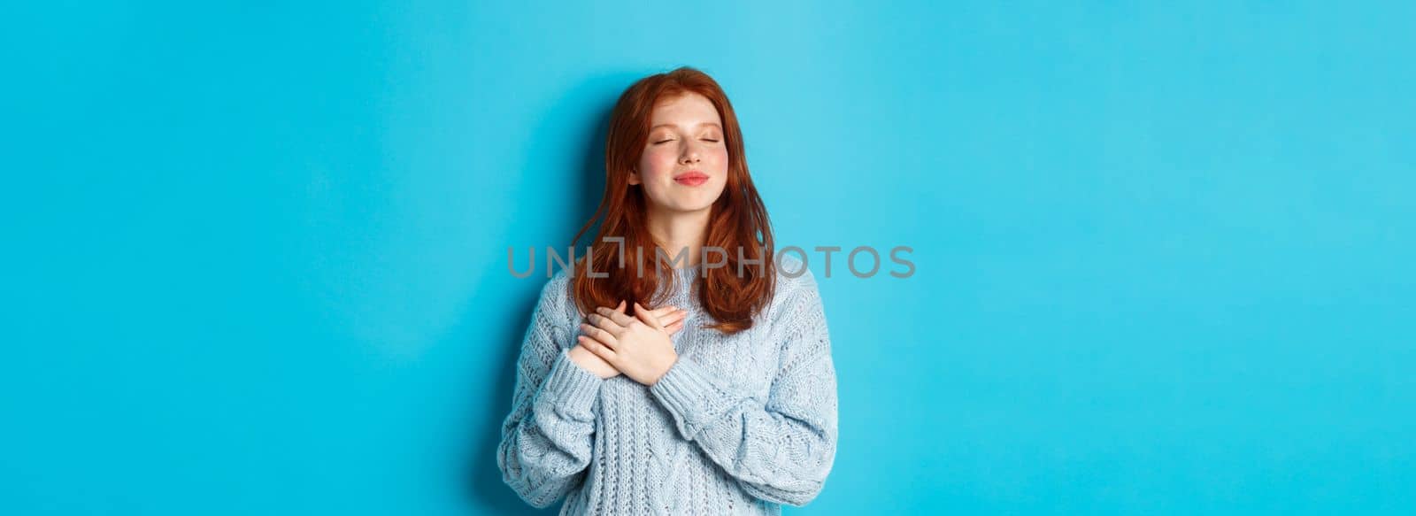 Nostalgic redhead girl in sweater dreaming, close eyes and holding hands on heart, remember something or daydreaming, standing over blue background.
