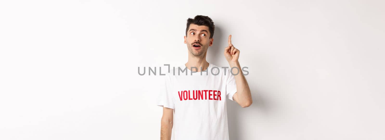 Handsome young man in volunteer t-shirt having an idea, raising finger and saying suggestion, white background.