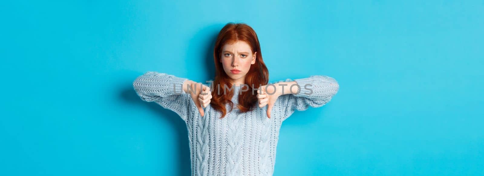 Disappointed redhead girl in sweater showing thumbs-down, judging bad product, disagree and dislike promo, standing over blue background.