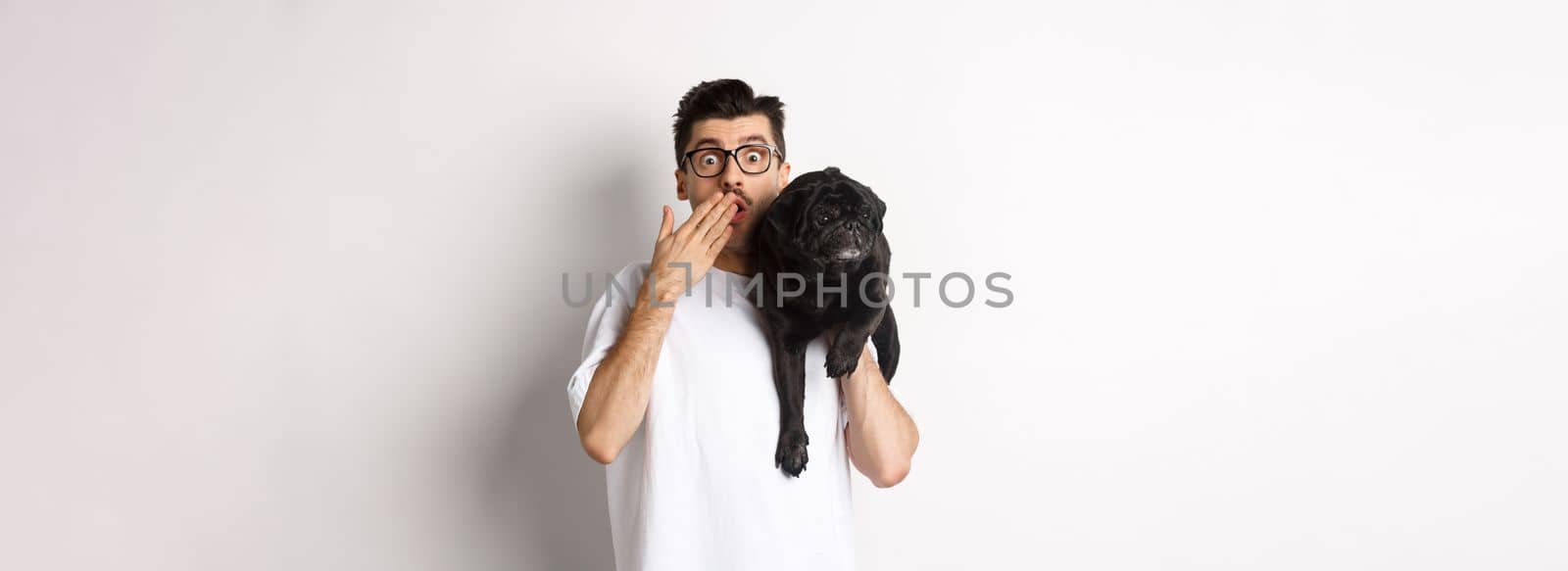 Shocked dog owner staring at camera, holding puppy on shoulder and gasping amazed. Handsome young man carry pug and reacting to promo advertisement, white background.