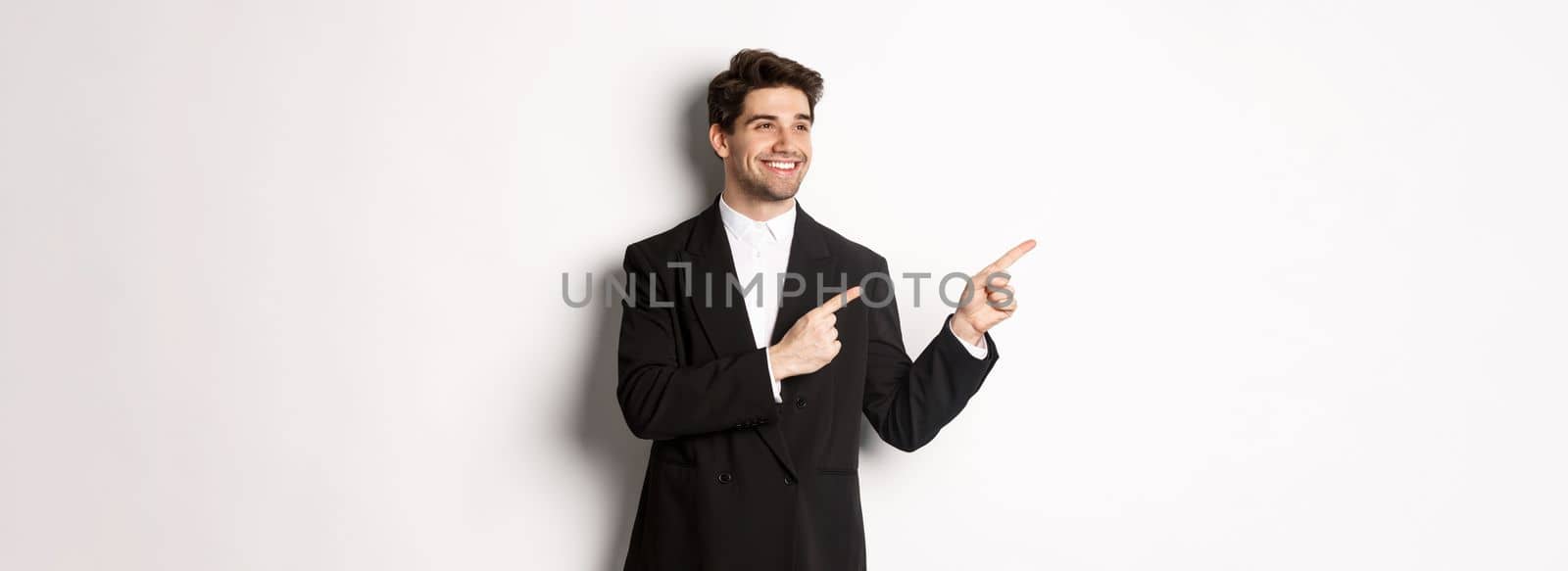 Portrait of successful handsome man in suit, pointing and looking left with pleased smile, showing promo banner, standing over white background.