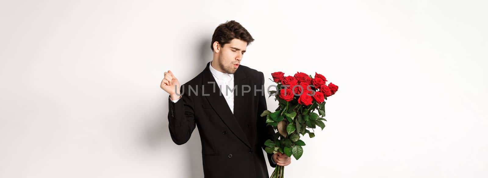 Image of elegant and sassy man in black suit, looking confident and holding bouquet of red roses, going on a romantic date, standing against white background by Benzoix