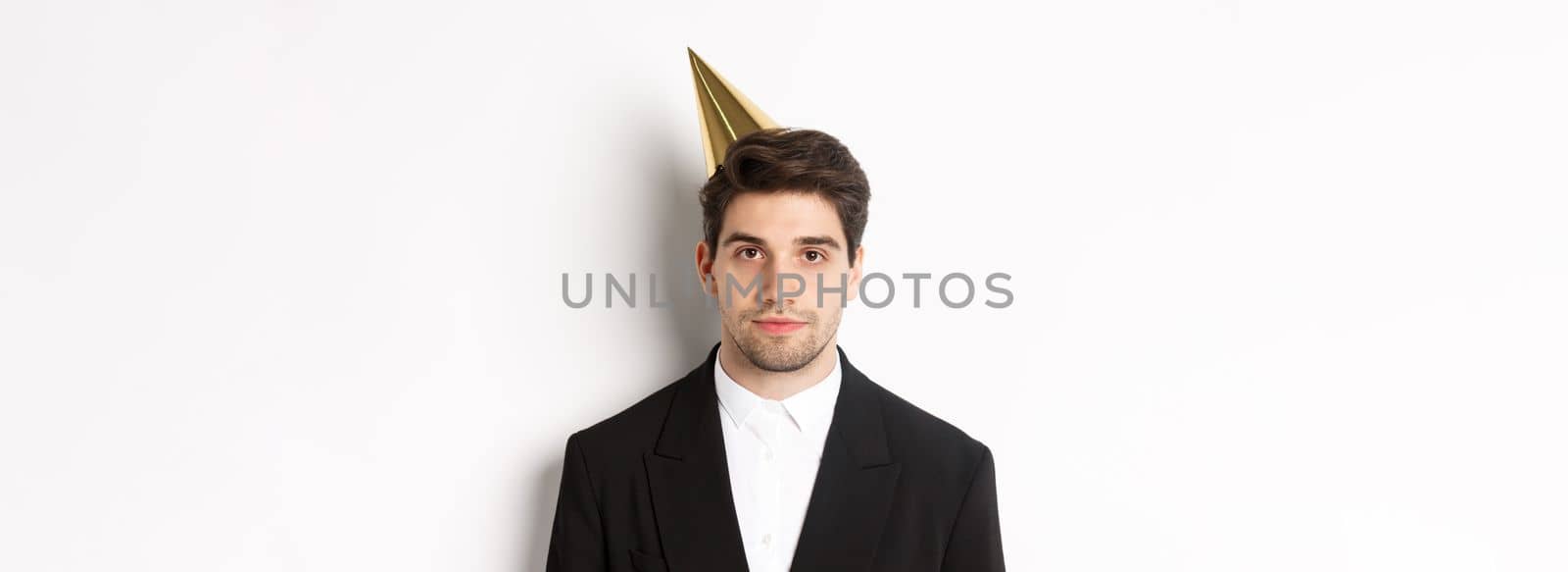 Close-up of handsome man in party hat and trendy suit, celebrating new year, standing against white background.