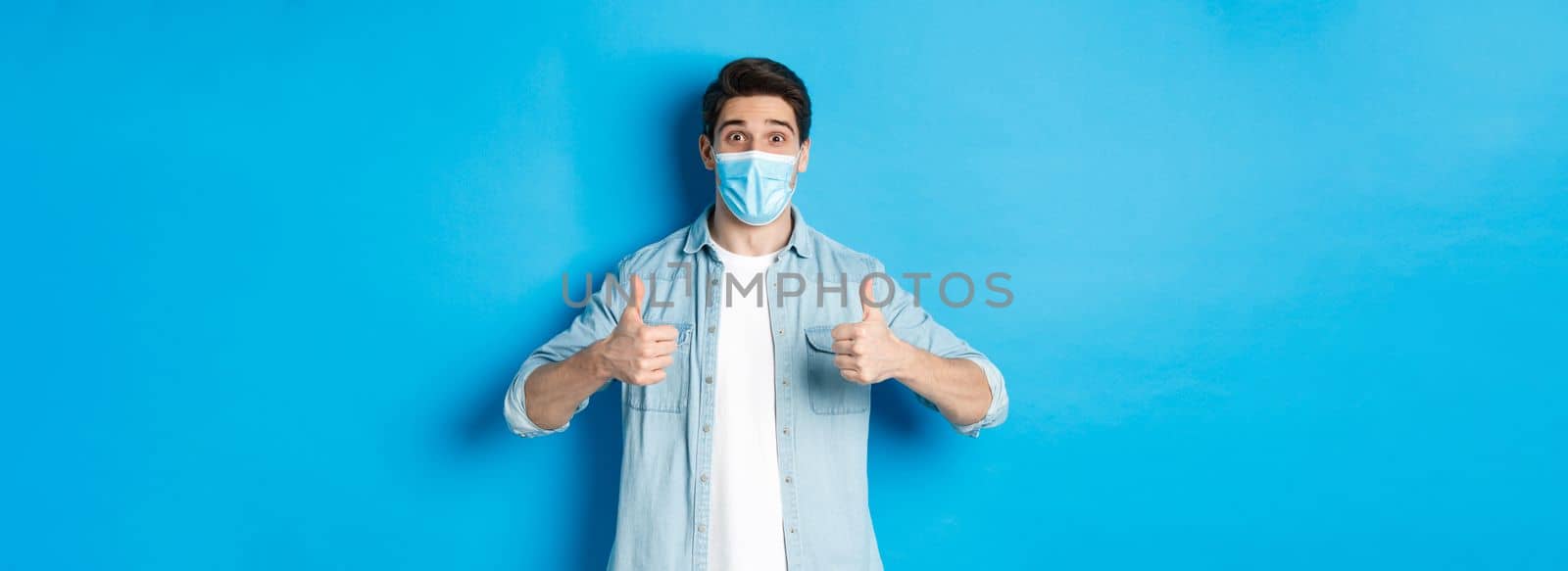 Concept of covid-19, pandemic and social distancing. Amazed guy in medical mask showing thumbs-up, recommending promo offer, standing against blue background.