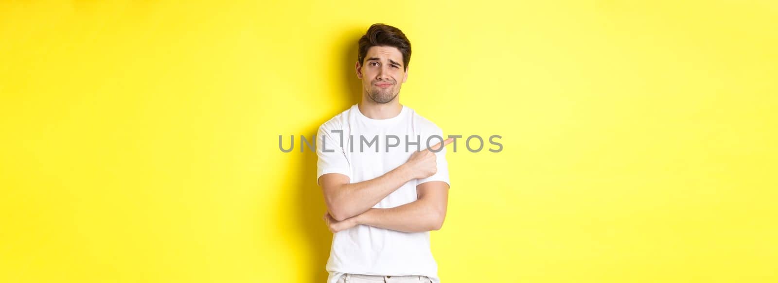 Skeptical and reluctant guy in white t-shirt, pointing finger left and smirking unamused, showing bad advertisement, standing over yellow background by Benzoix