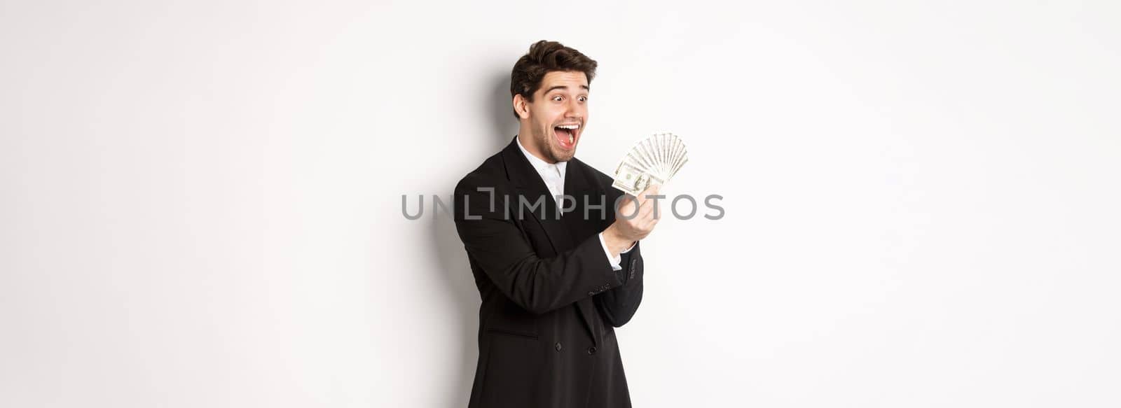 Image of excited handsome businessman in suit, looking amazed at money, winning cash, standing over white background.