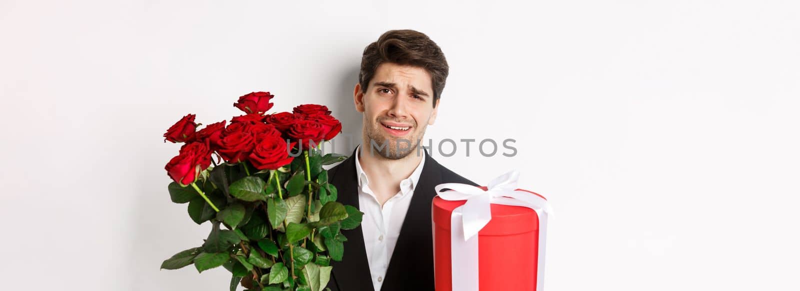 Close-up of skeptical man in suit, holding bouquet of red roses and a gift, standing reluctant against white background.