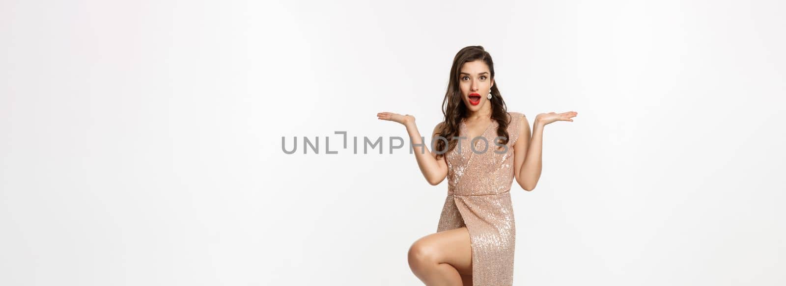 Christmas party and celebration concept. Attractive brunette model in elegant dress, showing two offers, raising hands and holding something on copy space, white background by Benzoix