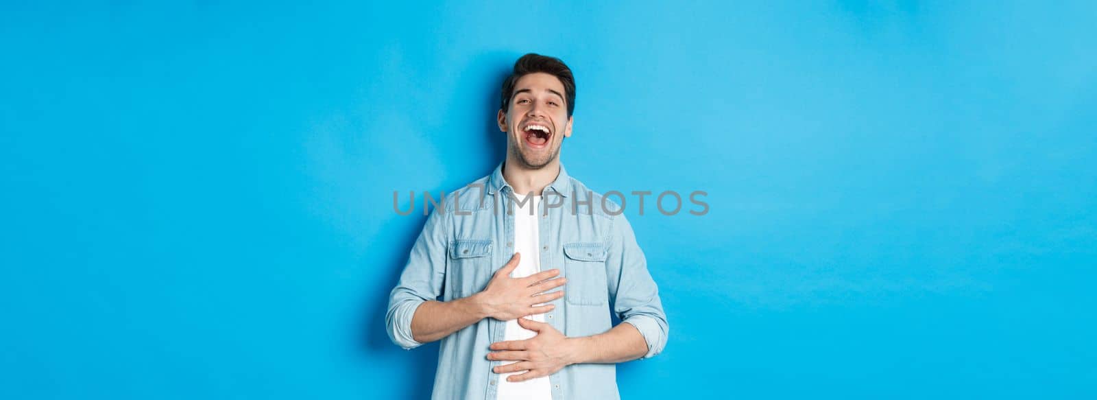 Happy handsome 30 year old man touching belly, laughing out loud and looking at something funny, standing against blue background.