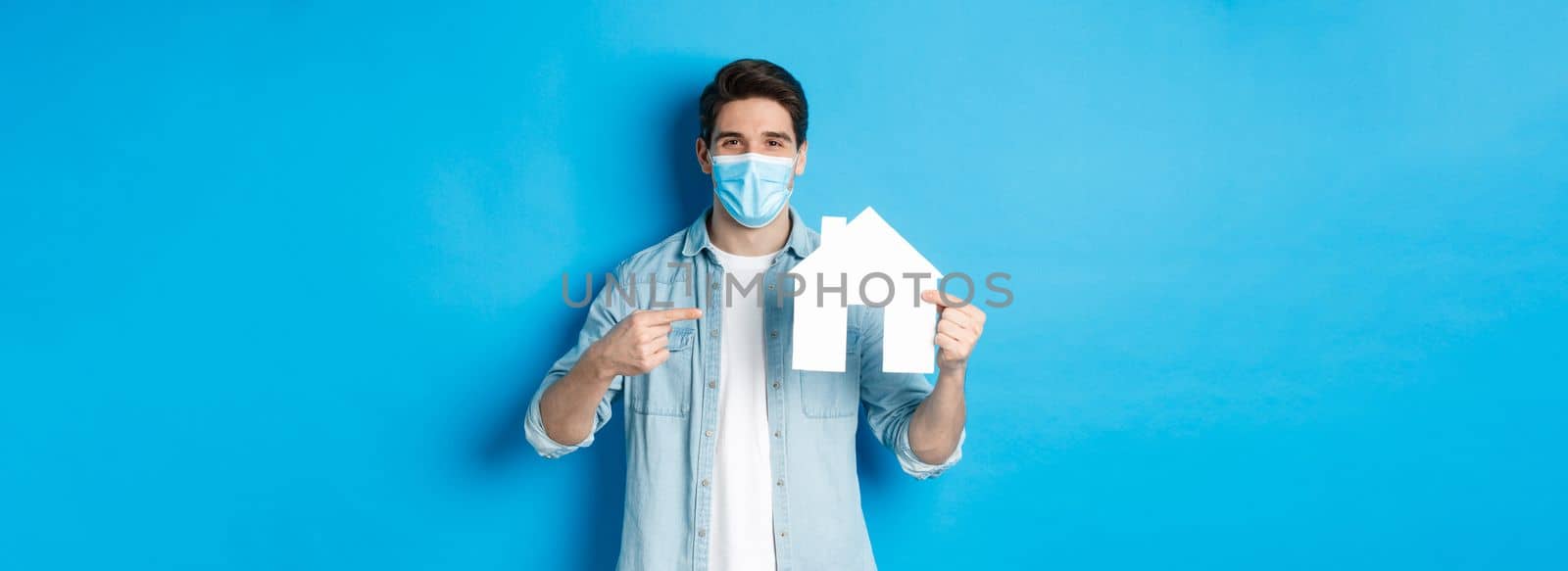 Concept of coronavirus, quarantine and social distancing. Young man searching aparment for rent, business loans, pointing at house model, wearing medical mask, blue background.