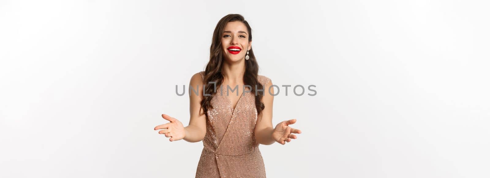 Attractive and elegent woman with red lipstick, wearing party dress, reaching for a gift, receiving Christmas present, smiling pleased, standing over white background.