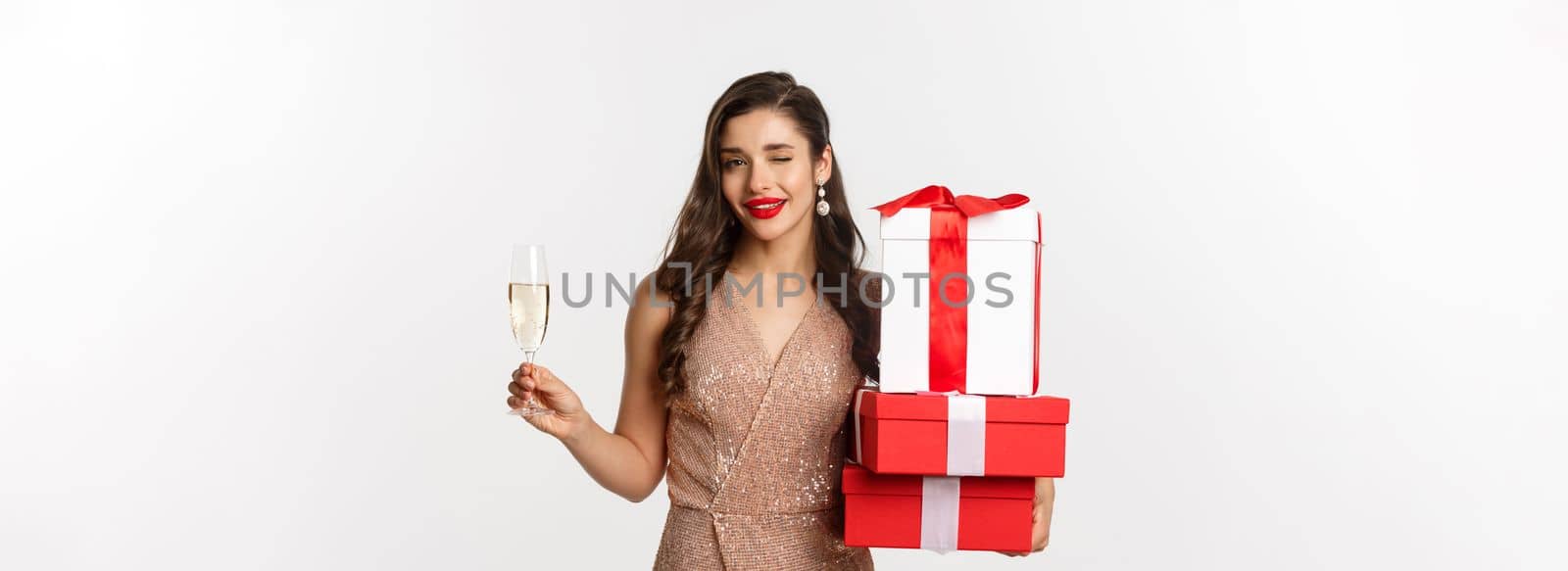 New Year, Christmas and celebration concept. Elegant woman in luxury dress and red lipstick, holding gifts and drinking champagne on party, standing over white background by Benzoix