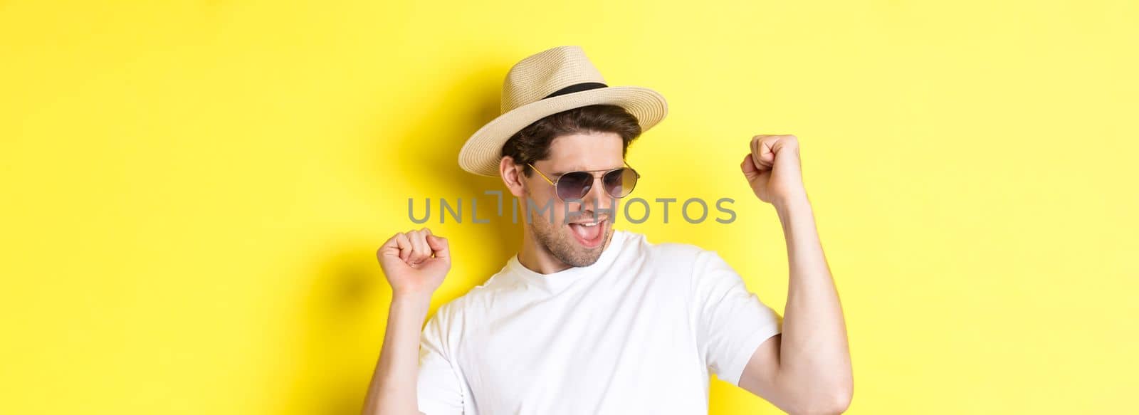 Concept of tourism and vacation. Close-up of man enjoying holidays on trip, dancing and pointing fingers sideways, wearing sunglasses with straw hat, yellow background.