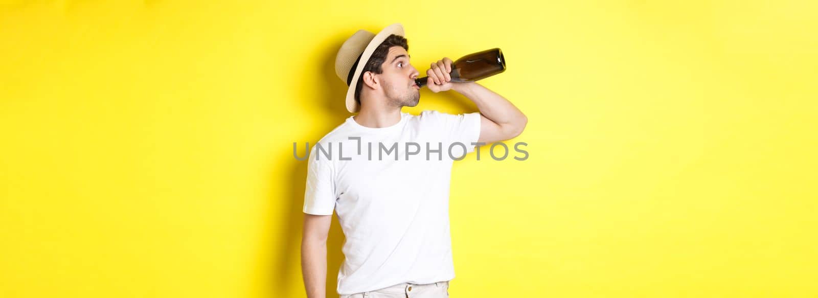 Concept of tourism and vacation. Man drinking wine from bottle on holidays, standing against yellow background. Copy space