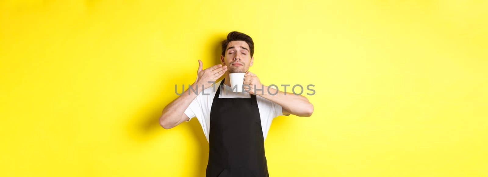 Barista enjoying smell of coffee in mug, standing pleased with eyes closed, wearing black apron.