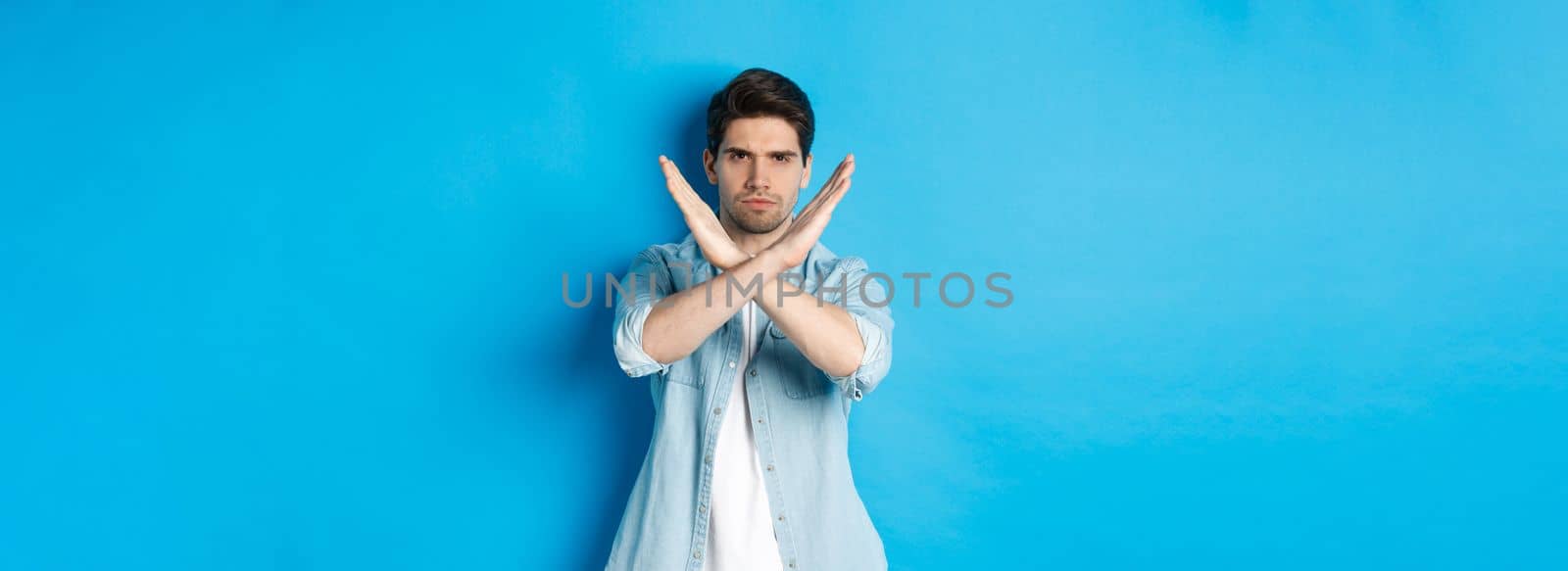 Displeased man frowning and saying no, making cross sign, tell to stop, standing against blue background.