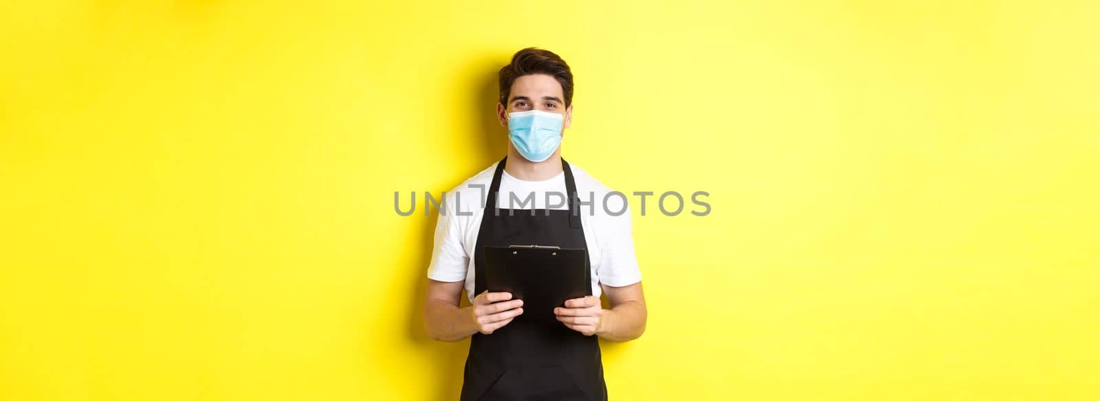 Concept of covid-19, small business and quarantine. Salesman in black apron and medical mask holding clipboard, working in shop, standing over yellow background.
