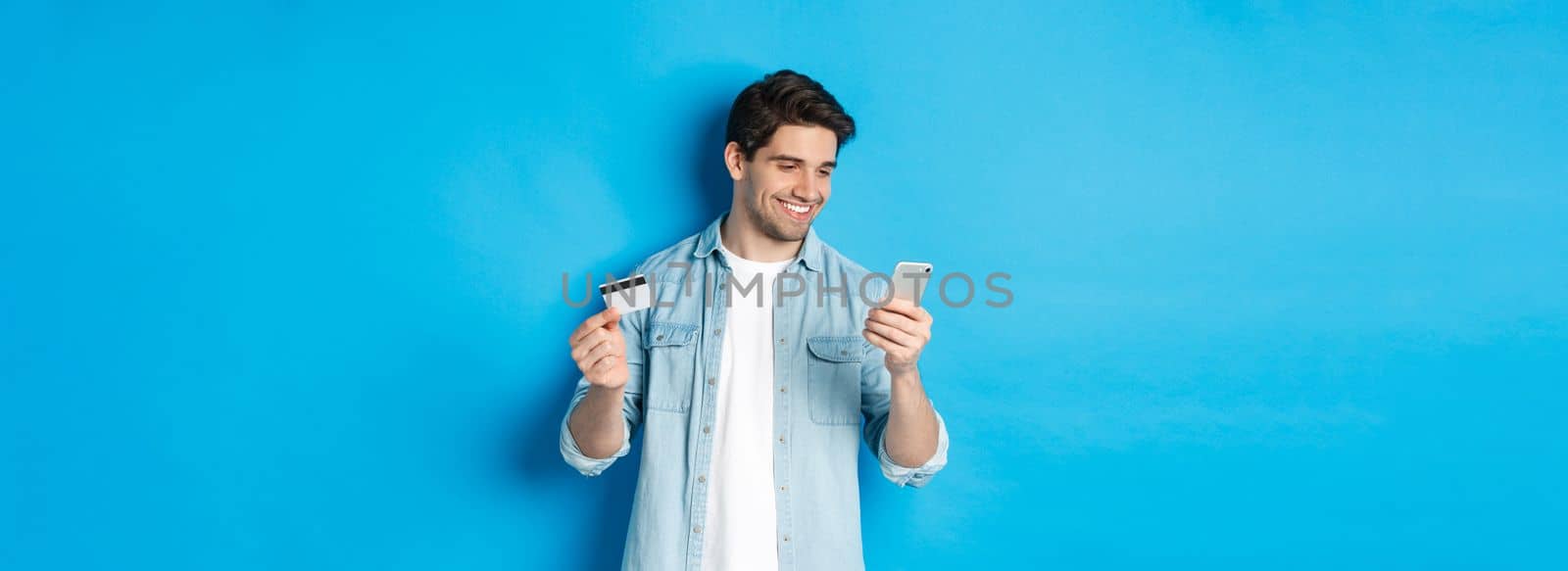 Young man shopping online with mobile application, holding smartphone and credit card, standing over blue background.