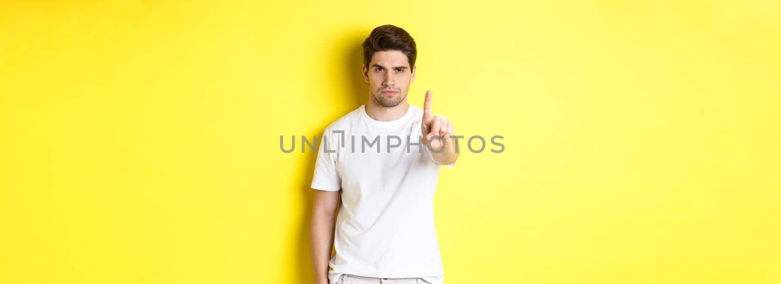 Confident man saying no, decline and prohibit something, showing one finger and frowning, standing over yellow background.