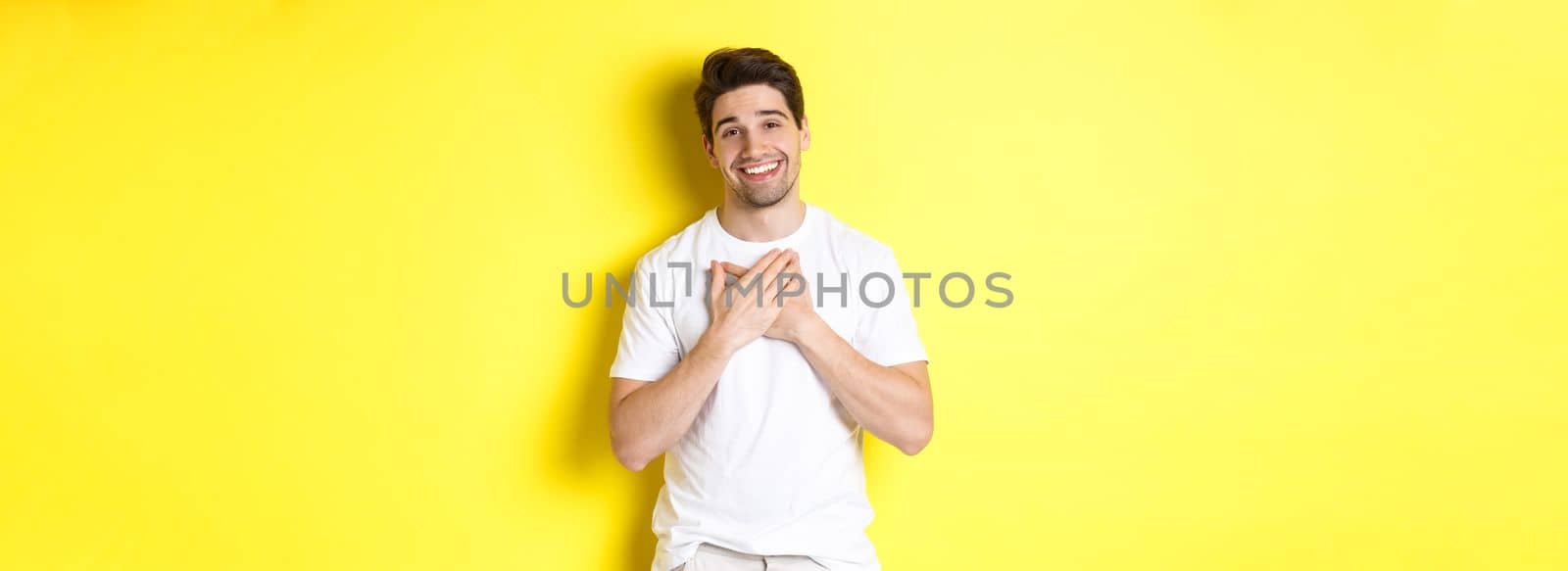 Image of grateful handsome guy in white t-shirt, holding hands on heart and smiling pleased, express gratitude, thanking for something, standing over yellow background.