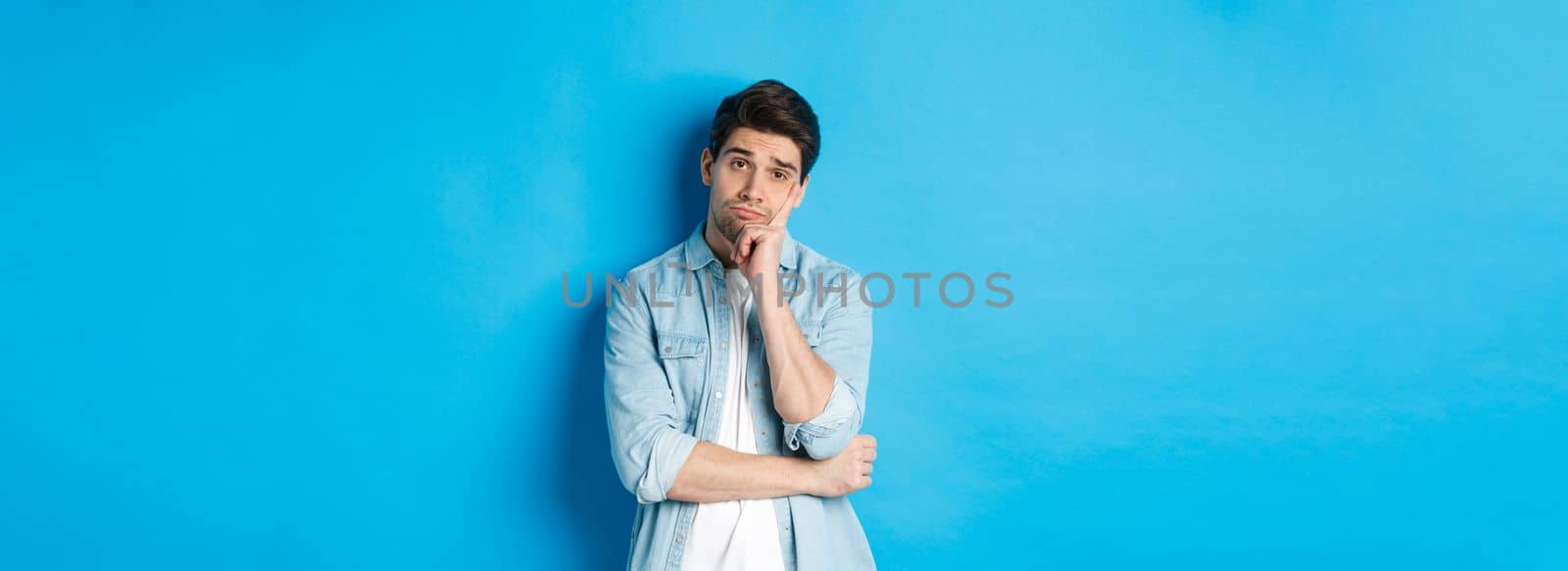 Unamused and bored man looking without interest at camera, standing against bllue background.