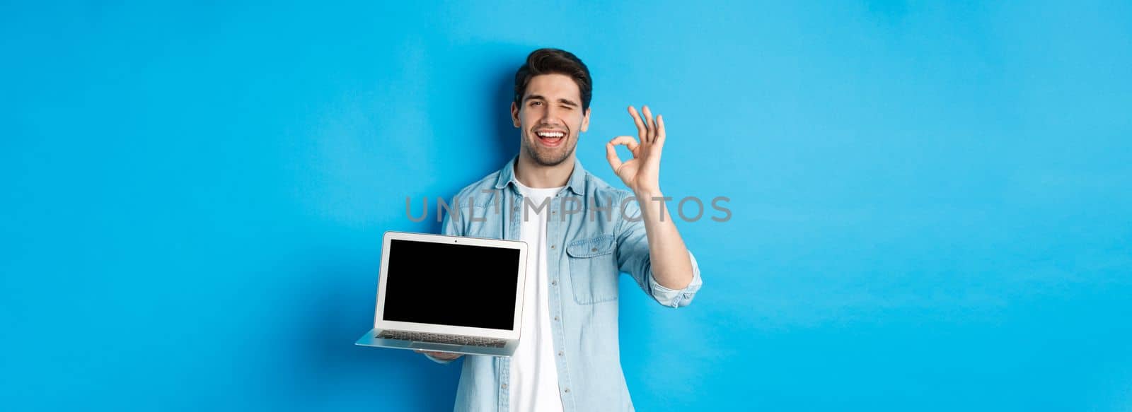 Young man showing laptop screen and okay sign, approve or like promo in internet, smiling satisfied, standing over blue background.