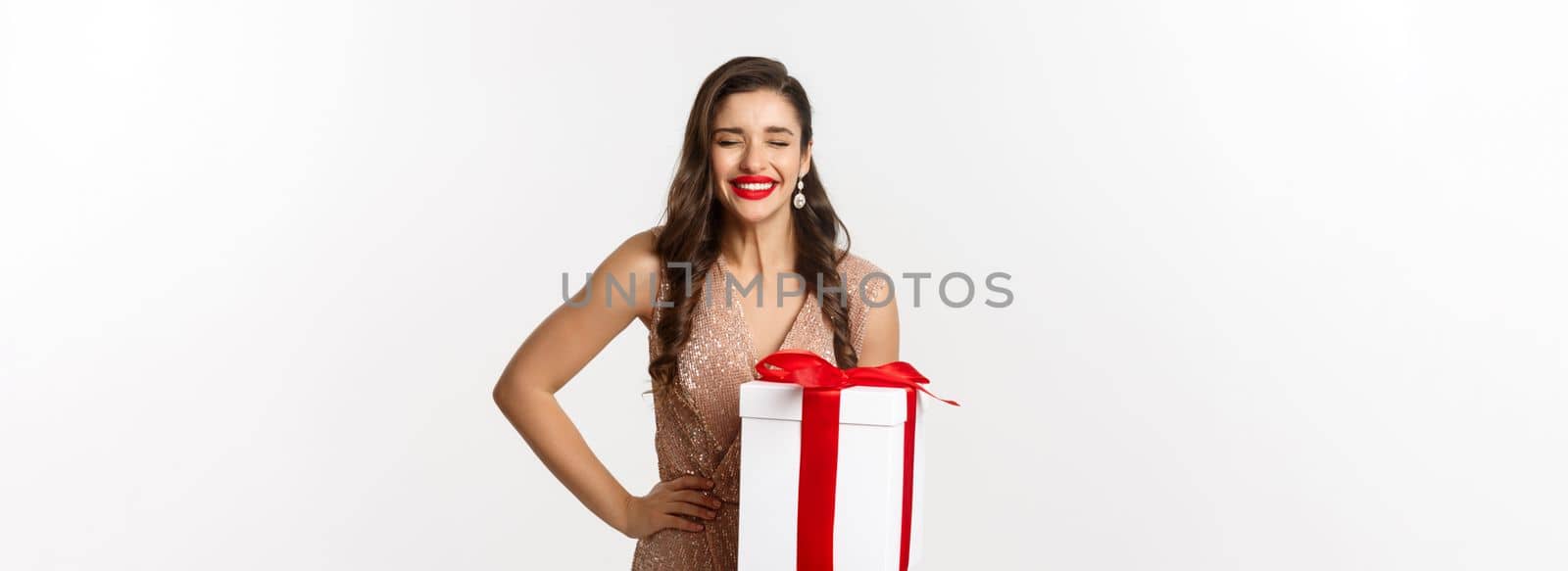 Merry Christmas. Image of attractive woman in luxury dress, celebrating winter holidays, holding gift and laughing from happiness, standing over white background.