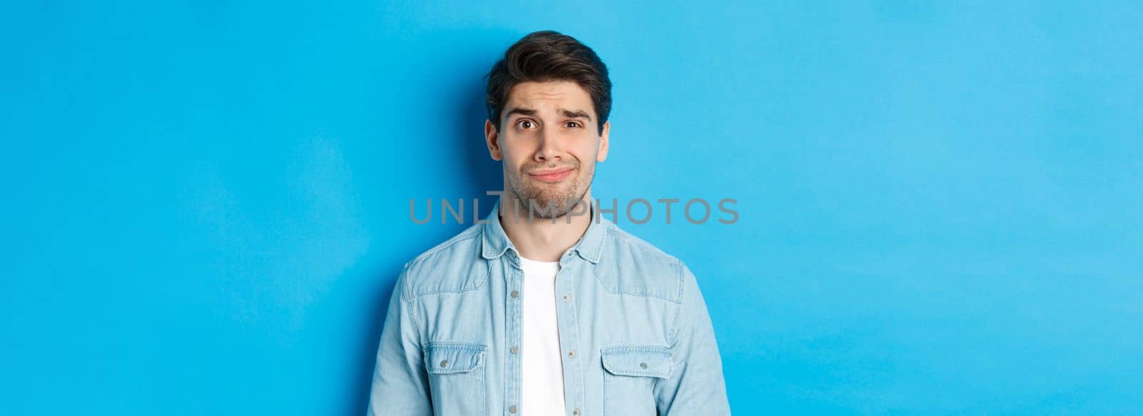 Close-up of skeptical and awkward guy smirking, feeling uncomfortable, standing over blue background.