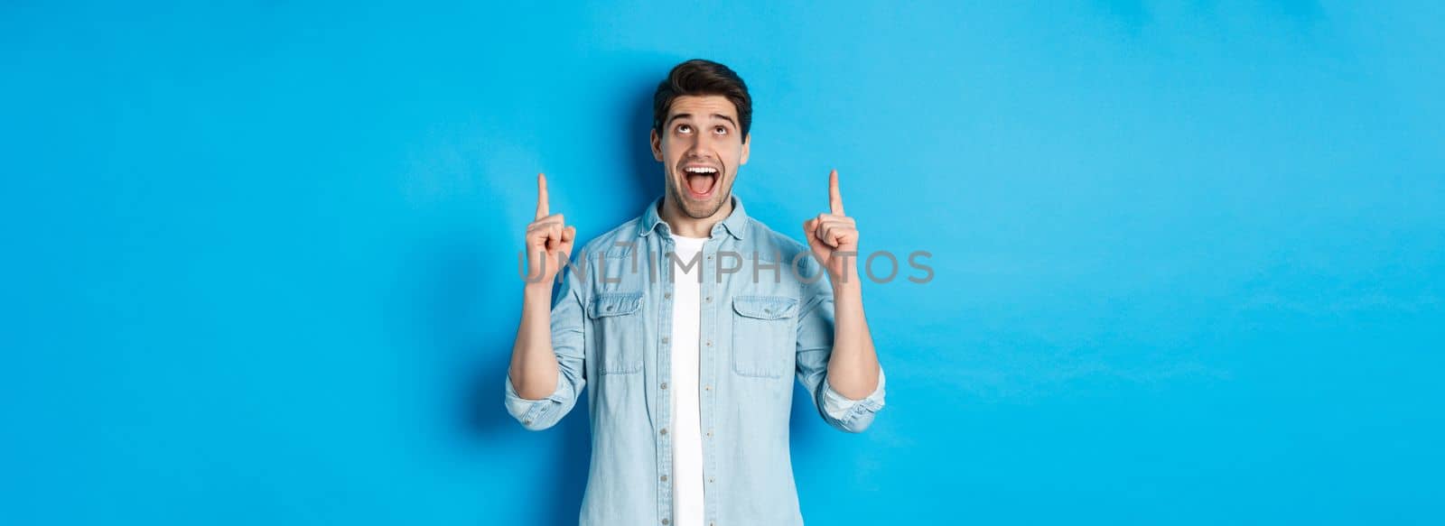 Excited handsome man pointing and looking up, showing advertisement, standing over blue background.