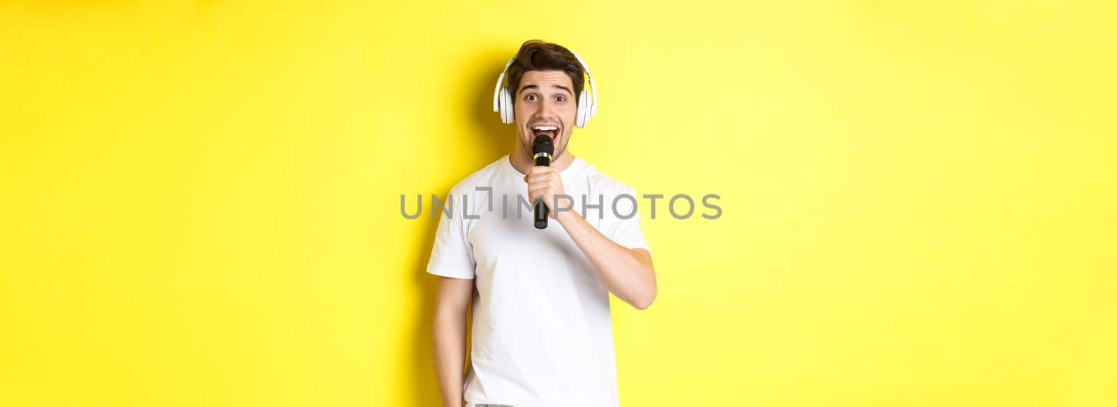 Man in headphones holding microphone, singing karaoke song, standing over yellow background in white clothes by Benzoix
