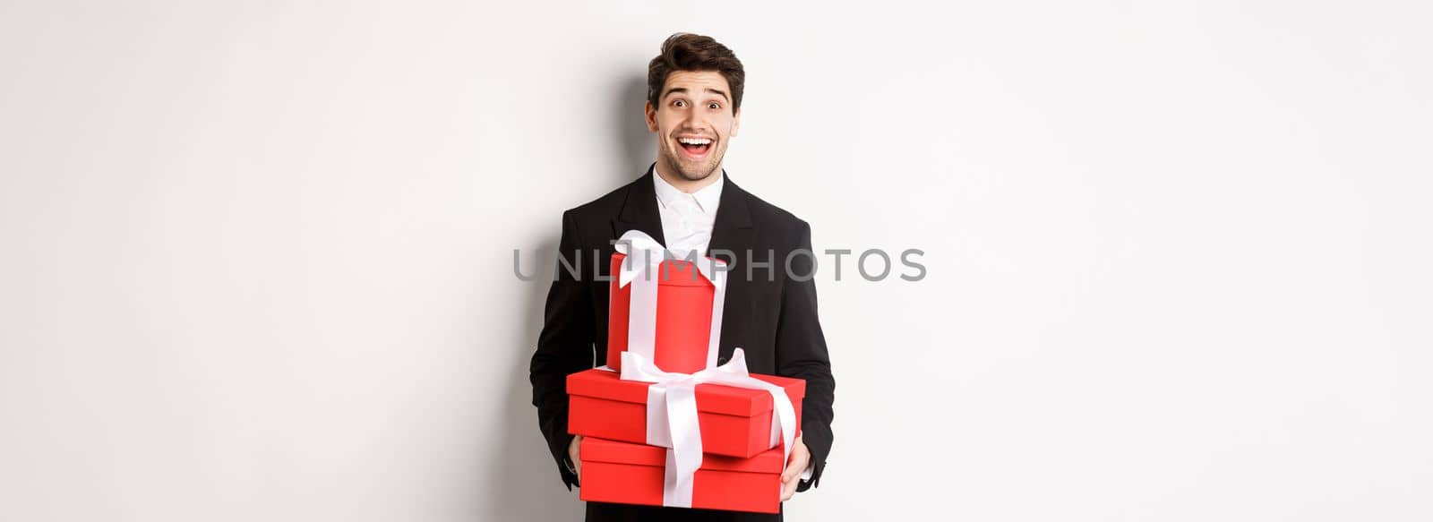 Concept of holidays, relationship and celebration. Handsome man in black suit bringing presents at new year party, holding gifts and smiling amused, standing against white background.
