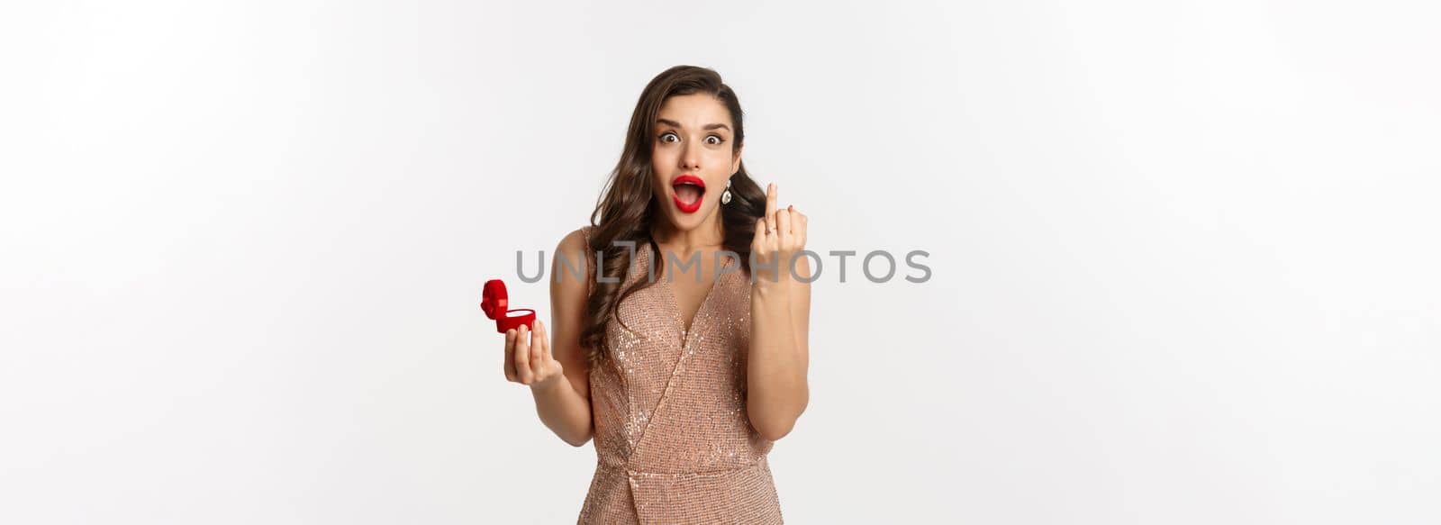 Excited young woman in glamour dress being engaged, showing finger with ring and looking happy, receive marriage proposal, standing over white background.