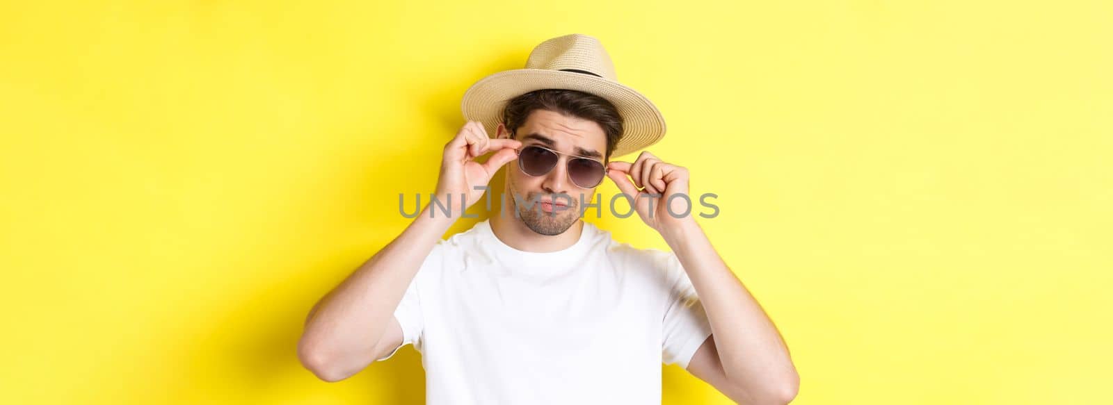 Concept of tourism and vacation. Close-up of cool tourist enjoying holidays on trip, wearing sunglasses with straw hat, yellow background.