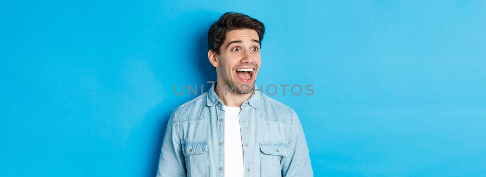 Close-up of handsome caucasian male model looking amazed left, checking out advertisement, standing over blue background.