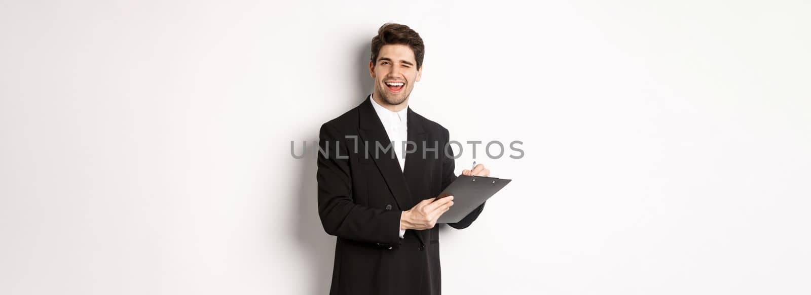 Image of handsome, successful boss in black suit, winking and smiling while signing documents, standing against white background.