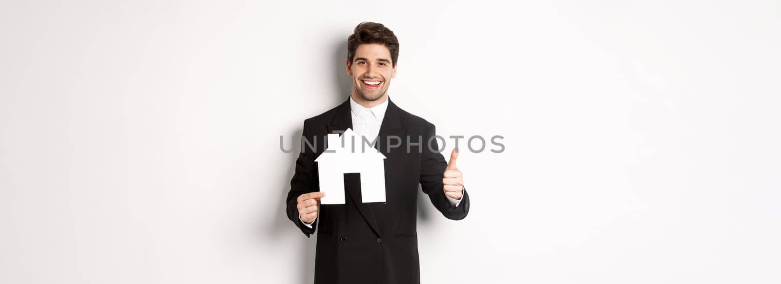 Portrait of confident real estate agent showing house maket and thumb-up, standing against white background by Benzoix