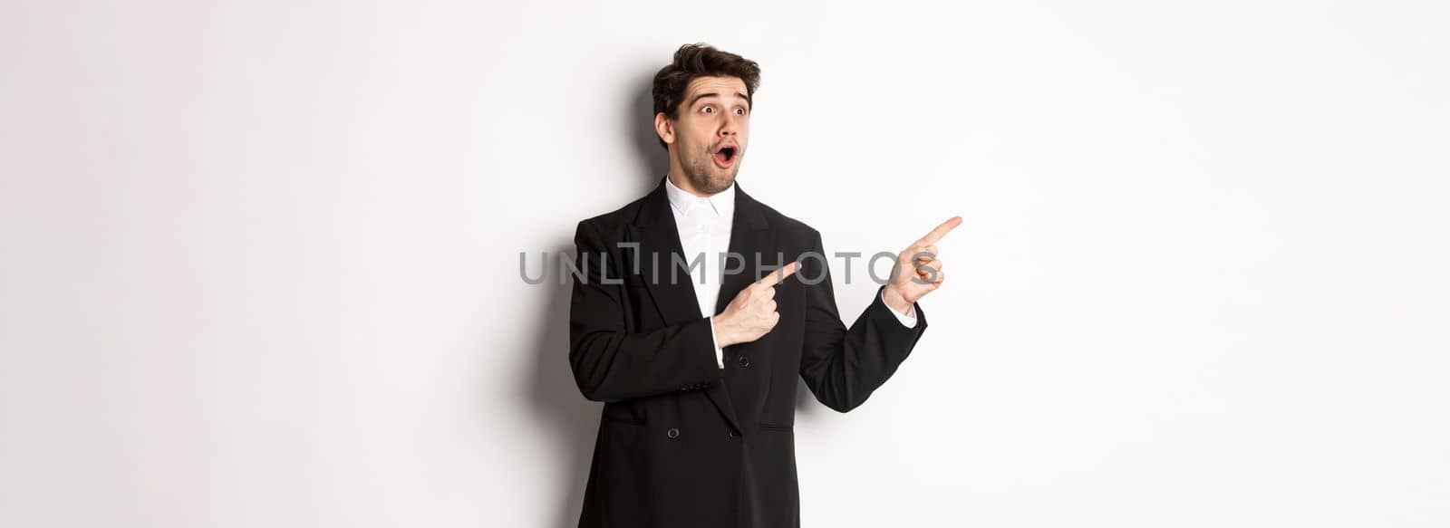 Impressed handsome man in party suit, looking at new year promo offer and pointing fingers left at banner, standing over white background by Benzoix