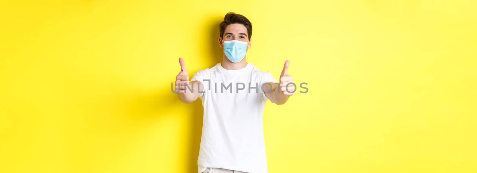 Concept of coronavirus, pandemic and social distancing. Confident man protecting himself from covid-19 with medical mask, showing thumbs up in approval, yellow background.