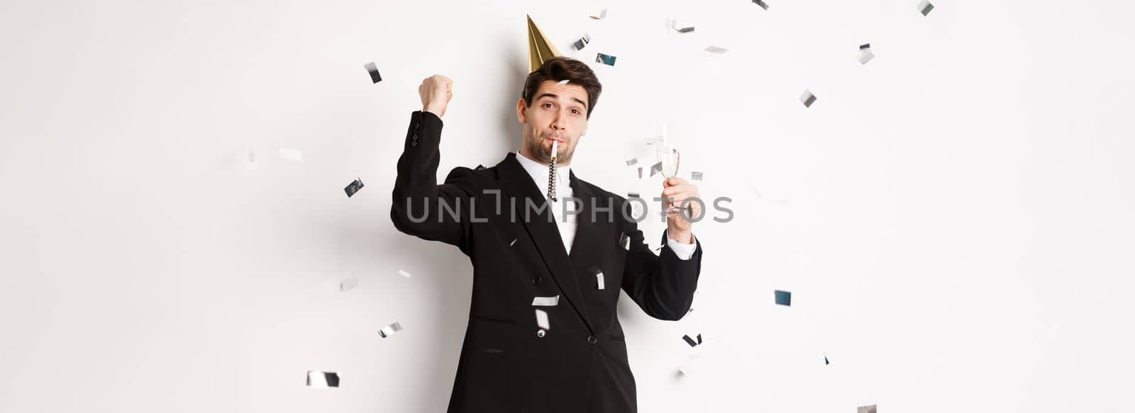 Handsome party guy in black suit having fun, celebrating new year, blowing whistle and drinking champagne while confetti falling, standing happy against white background.