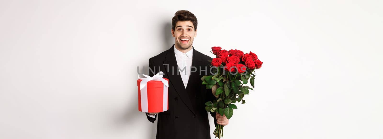 Concept of holidays, relationship and celebration. Image of handsome smiling guy in black suit, holding bouquet of red roses and giving present on new year, white background by Benzoix
