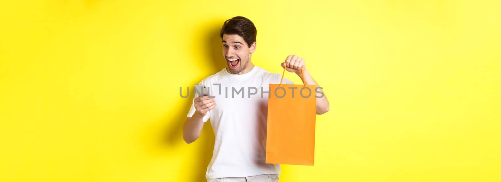 Concept of discounts, online banking and cashback. Surprised man showing shopping bag and looking happy at mobile screen, standing against yellow background.