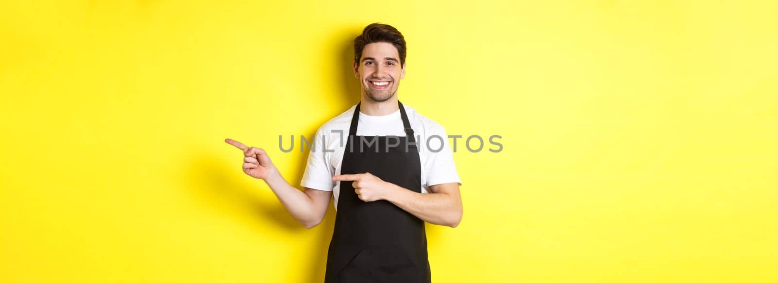 Friendly waiter pointing fingers right, showing your logo or promo offer, wearing black apron uniform, standing over yellow background,