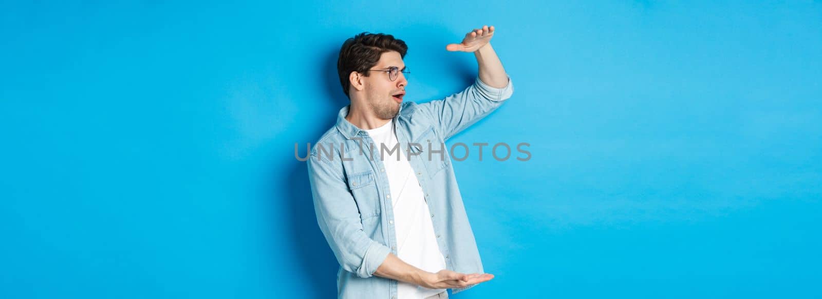 Excited handsome man showing big size object and looking amazed, standing over blue background.