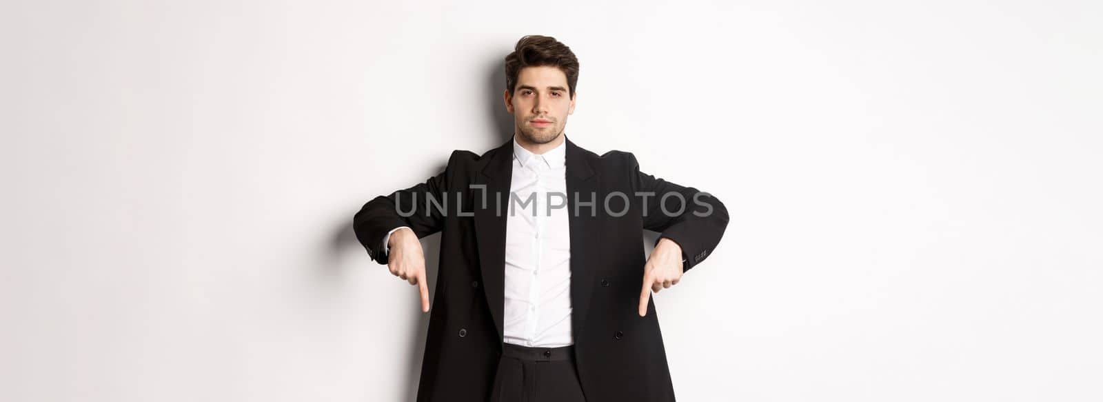 Image of handsome man dressed for formal party, wearing suit and pointing fingers down, showing advertisement or making announcement, standing over white background by Benzoix