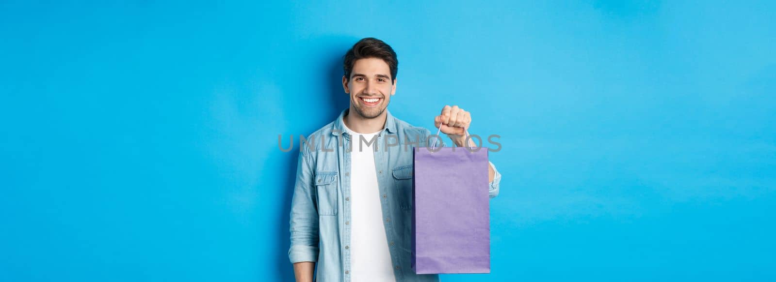 Concept of shopping, holidays and lifestyle. Young handsome man holding paper bag with present and smiling, standing over blue background by Benzoix